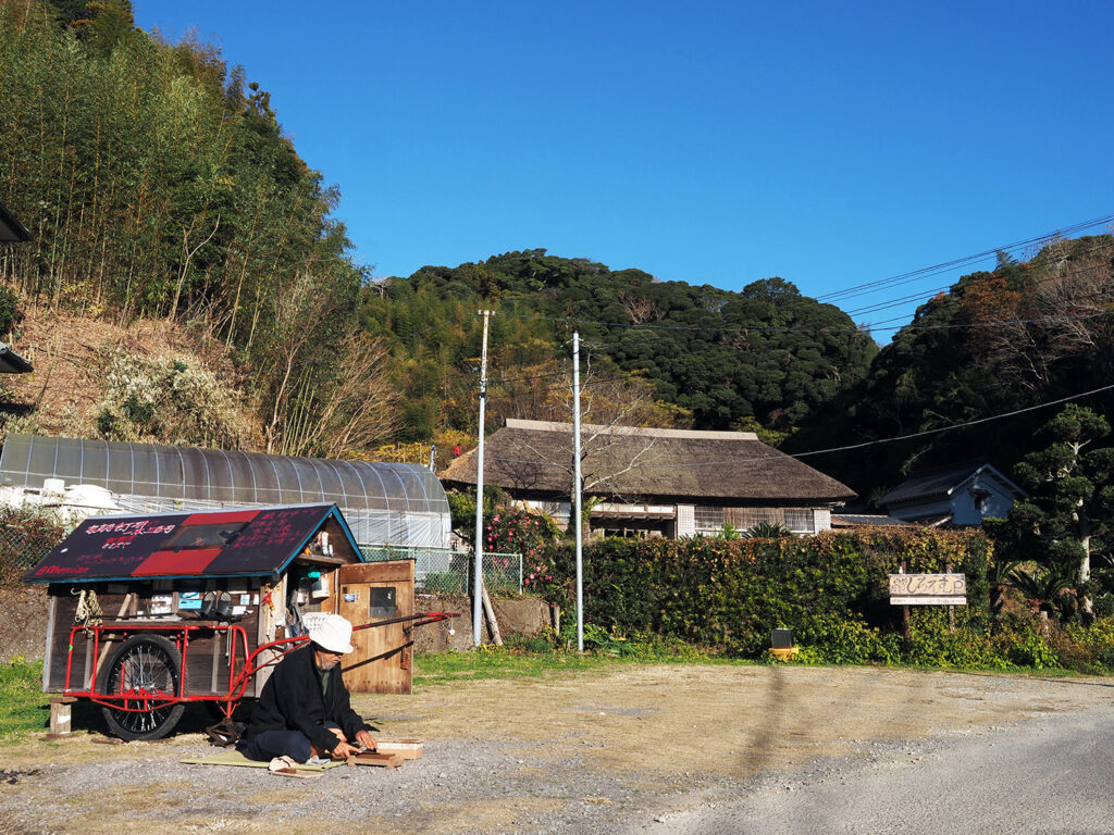 茅葺きの長屋門が見える駐車場で包丁を研ぐはるじさん